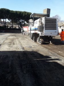 Macchina asfaltatrice e camion su strada sterrata.